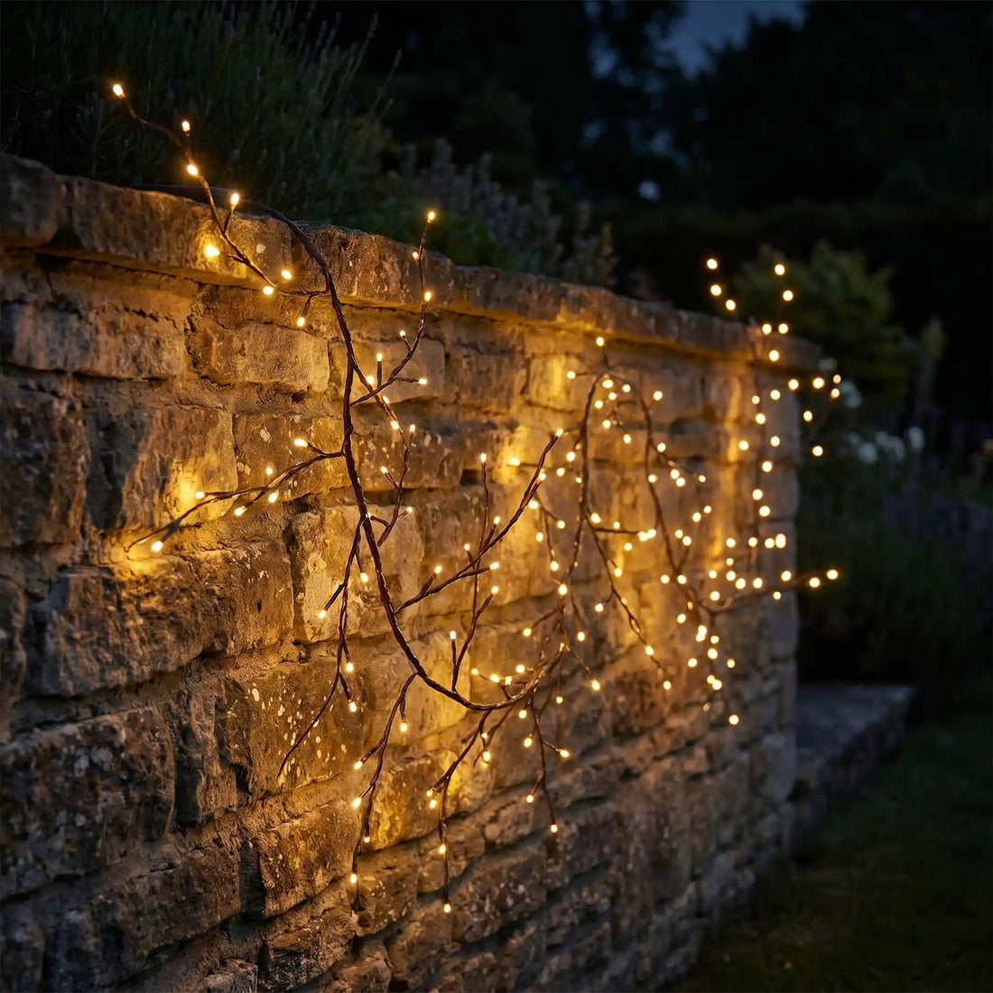 Guirlande lumineuse extérieur solaire style branche qui serpente sur un mur en pierre la nuit, lumière ambrée chaude, jardin soigné