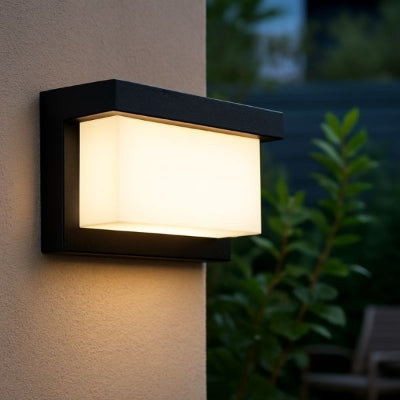 Gros plan d’une applique murale extérieure LED rectangulaire noire diffusant une lumière blanche froide, fixée sur un mur en crépi clair avec plantes vertes en arrière-plan.