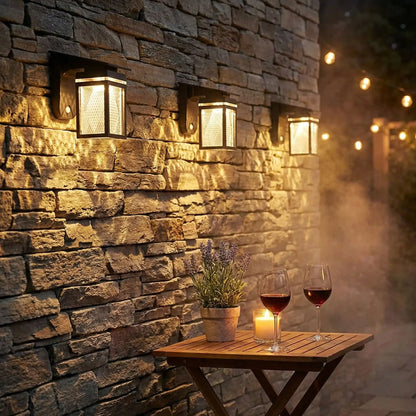applique solaire exterieur blanc chaud sur mur en pierres naturelles dans jardin, table  en boisavec deux verres de vin, une plante et une bougie allumée. Guirlande en arrière plan