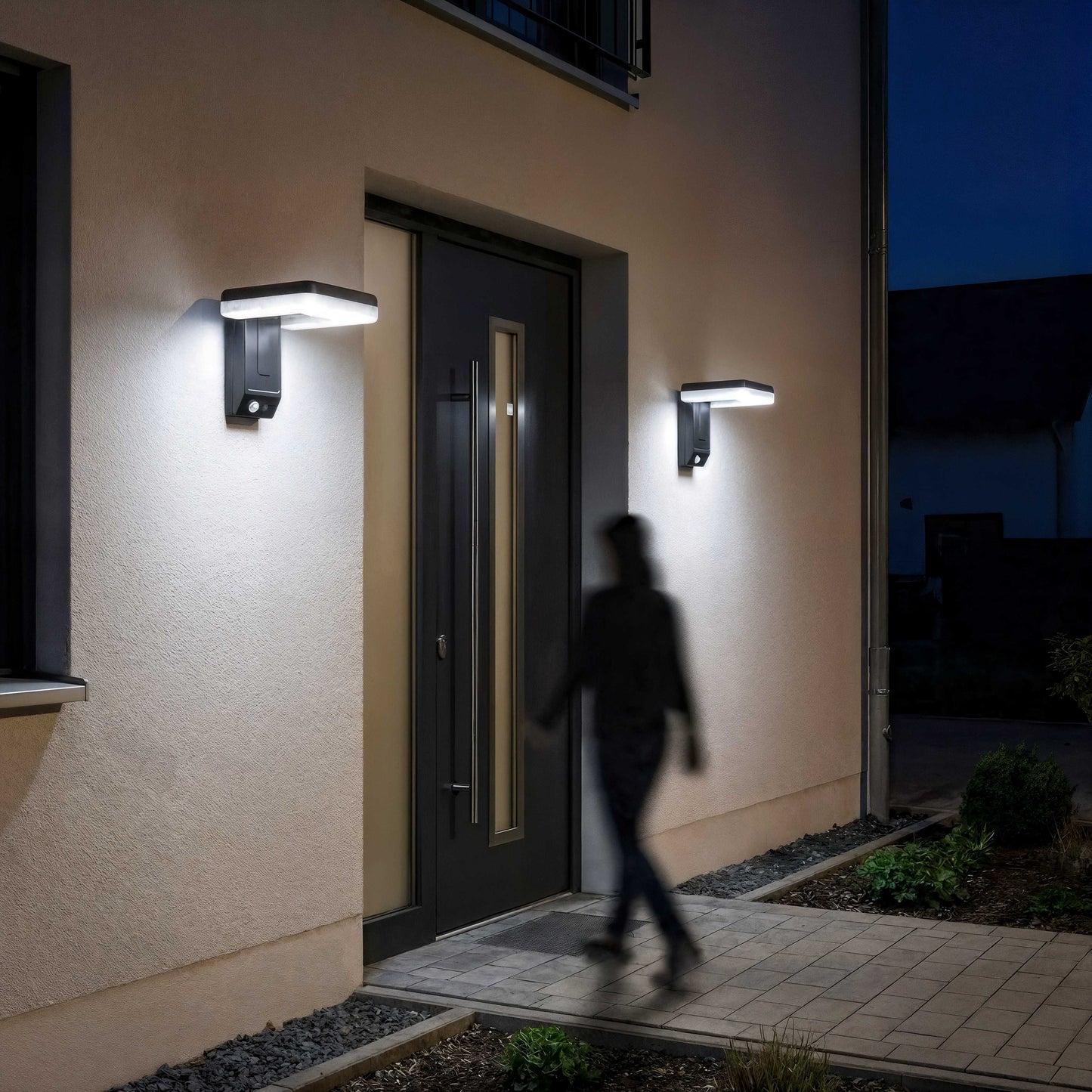 appliques exterieures solaires exterieures allumées à l'entrée d'une maison contemporaine et une silhouette floue de femme qui rentre dans la maison