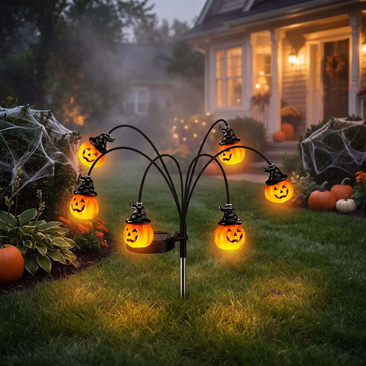Citrouille lumineuse solaire Halloween à 6 têtes et chapeaux de sorcière plantée dans la pelouse devant une maison décorée, à la tombée de la nuit