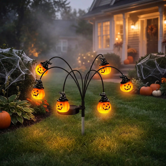 Citrouille pour halloween solaire à 6 têtes et chapeaux de sorcière plantée dans la pelouse devant une maison décorée, à la tombée de la nuit