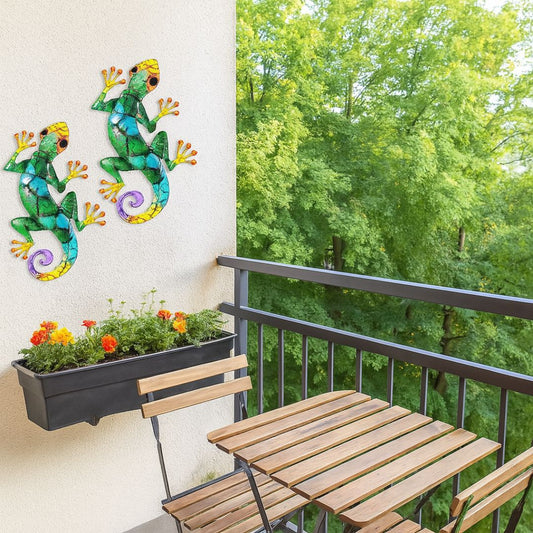 Deux sculptures en lézards muraux colorés décorent un balcon avec jardinière fleurie, table et chaises en bois, fond arboré.