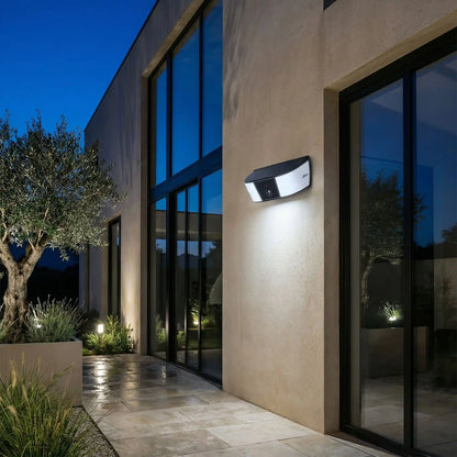 eclairage exterieur solaire avec detecteur sur devanture maison ontemporaine avec grandes baies vitrées, terrasse, olivier et plantes décoratives au crépuscule