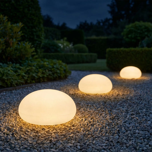 Galet solaire lumineux disposé en perspective sur une allée de graviers dans un jardin soigné la nuit, lumière ambrée chaude et douce